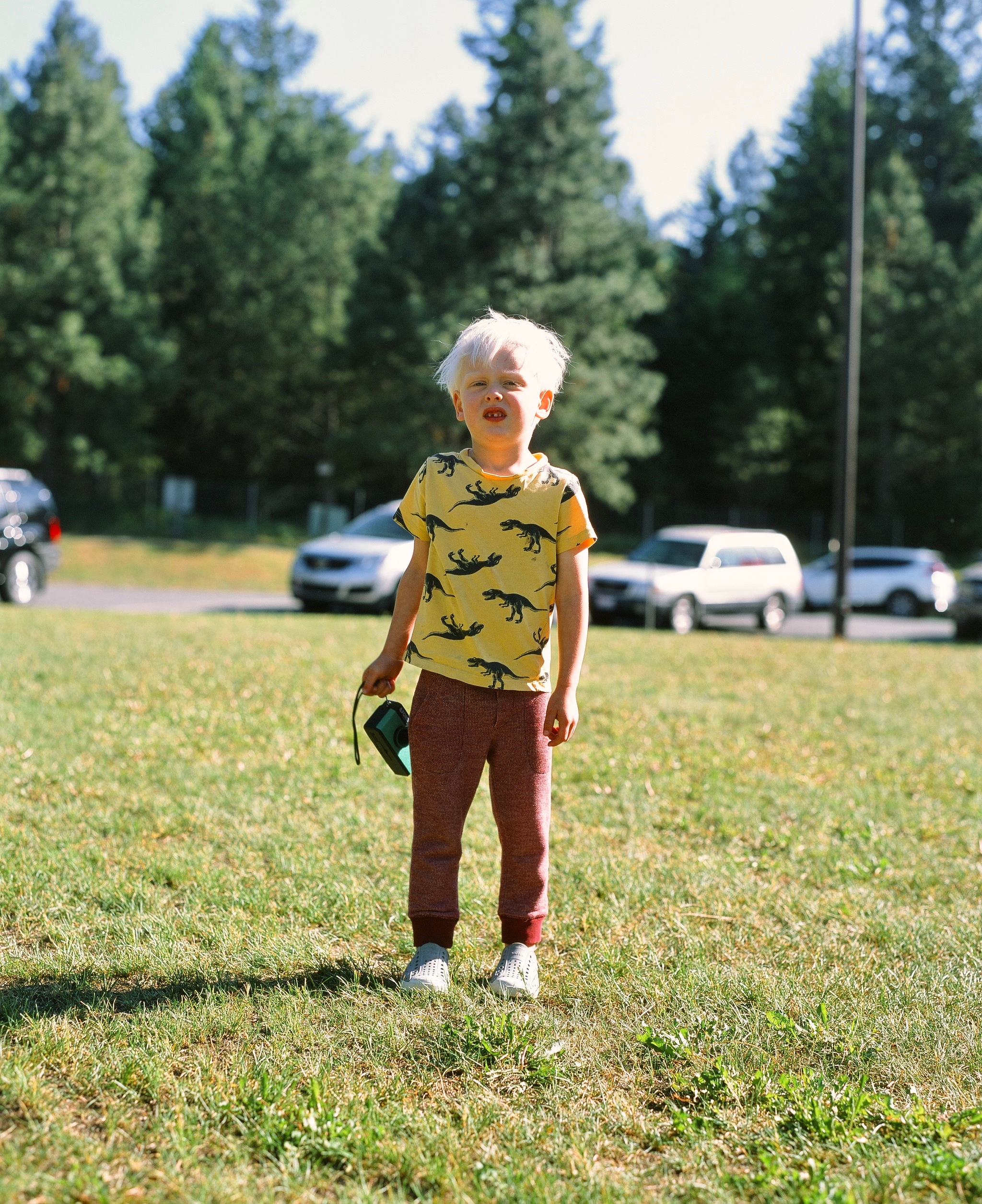 Young boy with white hair looking discheveled and tired, standing in the sun, holding a film camera.